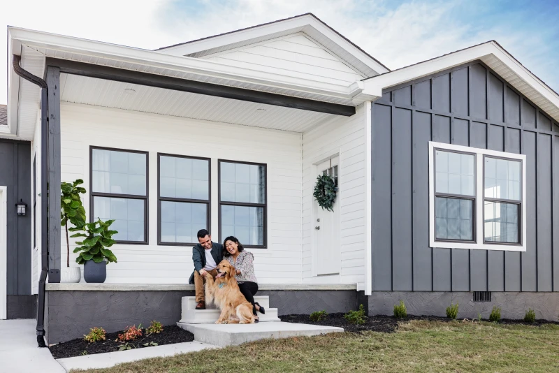 Young couple with a dog sit on the front step of a Clayton CrossMod home with a porch and white and gray siding