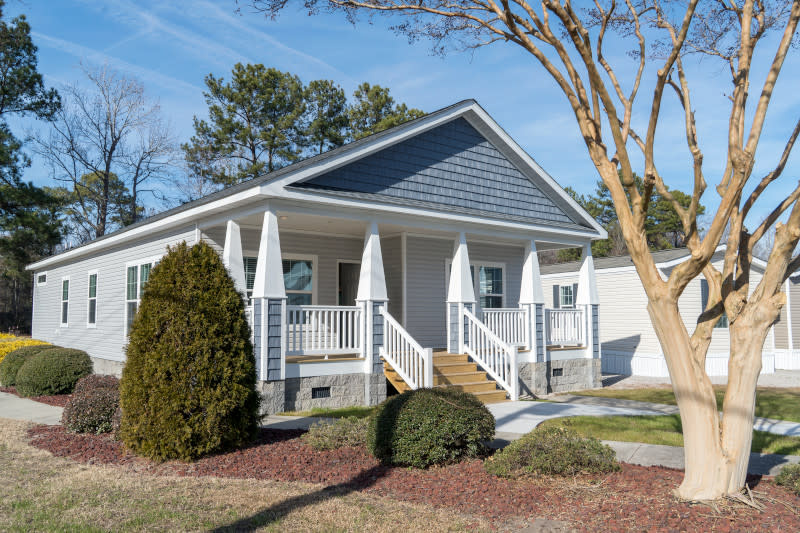 The gray, white and blue 2466 Oakwood Mod has a craftsman-style covered porch with ventilated brick skirting and a landscaped yard with a large tree in the front.