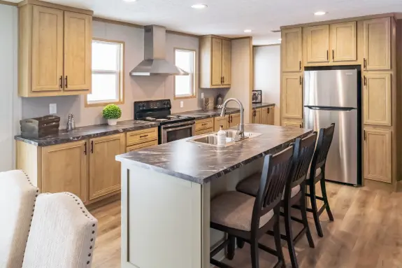 The [model name]'s kitchen has surrounding cabinets around the fridge and an island with marble-style counters.