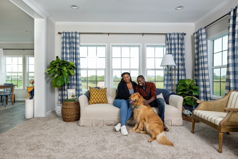  Couple sitting on couch petting a dog. Room has gingham blue and white curtains, fake trees, and wicker decor.