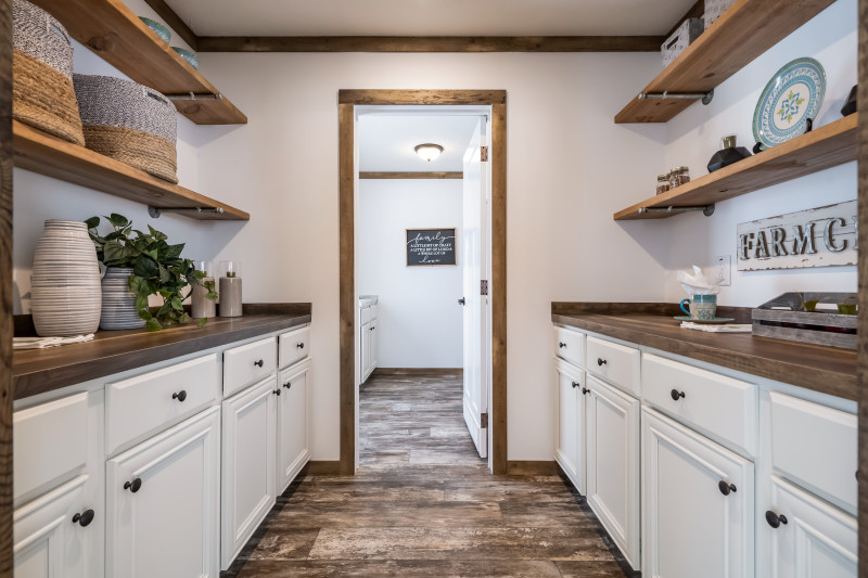 The pantry of a manufactured home kitchen with white cabinets, wood style counters and open shelves with farmhouse decor, leading into a utility room.