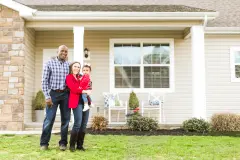 A young couple and their baby standing in front of their home.