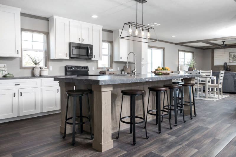 Manufactured home kitchen with white cabinets, gray wood-style flooring, metal chandelier and long, tan kitchen island with metal stools, and dining and living areas visible in the distance.