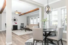 A farmhouse-style living room with rustic wall décor, a beige couch, rocking chairs and wood ceiling beams connects with a dining room with double windows, metal chandelier and a white and dark wood dining set with a bouquet in a glass jar on display.