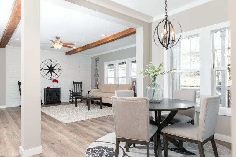 A farmhouse-style living room with rustic wall décor, a beige couch, rocking chairs and wood ceiling beams connects with a dining room with double windows, metal chandelier and a white and dark wood dining set with a bouquet in a glass jar on display.