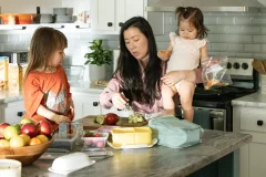 Woman holds a toddler while packing a lunch at a kitchen island, while another child sits nearby.