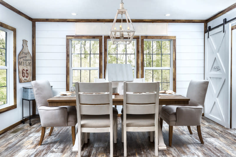 Dining room of a manufactured home featuring shiplap walls, wood trim, 3 windows, chandelier, wooden table, beige chairs and white sliding barn door.