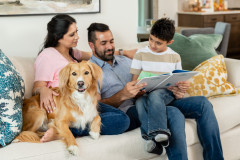 A family is gathered on a white couch with colorful pillows. They are sitting with their dog and looking through a book. 
