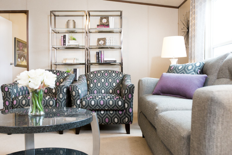 Two fun patterned dark gray, purple, and green chairs are sitting in a living room with a gray couch to the side. There’s a coffee table with white flowers in a vase on it. Sunlight fills the room. Plus, there are two silver rimmed bookshelves in the back of the photo view.