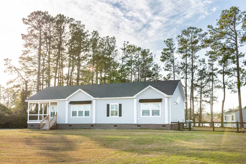 Exterior of manufactured home with side porch, gray siding and roof, brick skirting and tall trees behind it.