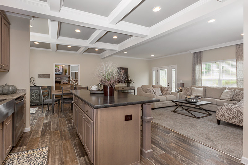 The open kitchen, dining and living room of the Kennesaw manufactured home with beige decor, island and coffered ceilings.