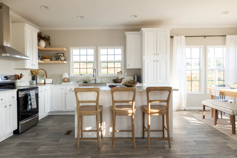 Kitchen of a Clayton manufactured home with white cabinets and an island with three chairs, weathered wood flooring and a dining room off to the side.