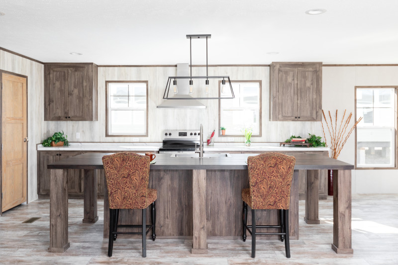 Kitchen in a manufactured home with large island with chairs pulled up to it. Kitchen is brown and white with a modern, industrial light fixture hanging over the island.