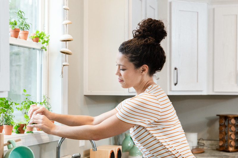 A woman with a striped shirt picks herbs in small clay pots on the windowsill over a sink faucet.