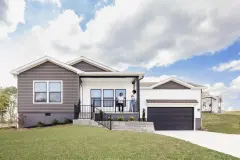 A manufactured home with white and gray siding, a garage and driveway and two people on the porch.