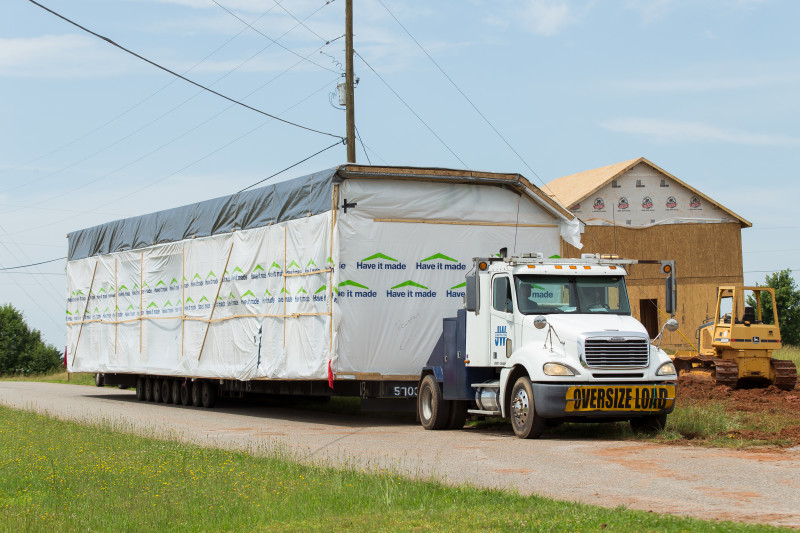 A section of a manufactured home is transported on a gravel road the back of a large truck to the final home site, with another home being built in the background.