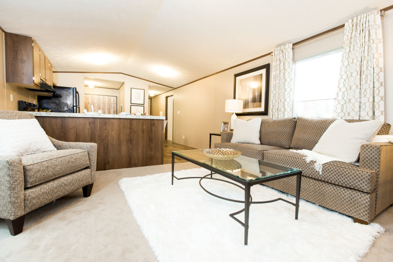 View is of a manufactured home living room with the kitchen to the left. The living room has a brown couch, tan chair, and white rug with a glass plated table on top of it.