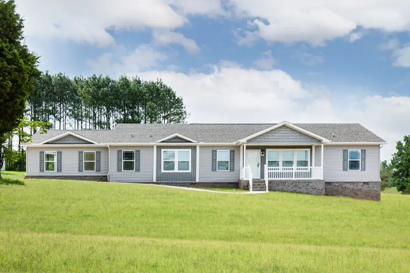 Exterior of a manufactured home from Clayton with gray siding, a gray roof, brick skirting and white trim, with a large grassy yard and trees and blue sky in the background.