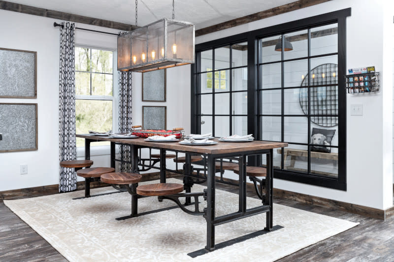 Dining room of a manufactured home with rustic metal chandelier, window and dining table with attached seats, with a paned glass wall showing the foyer behind.