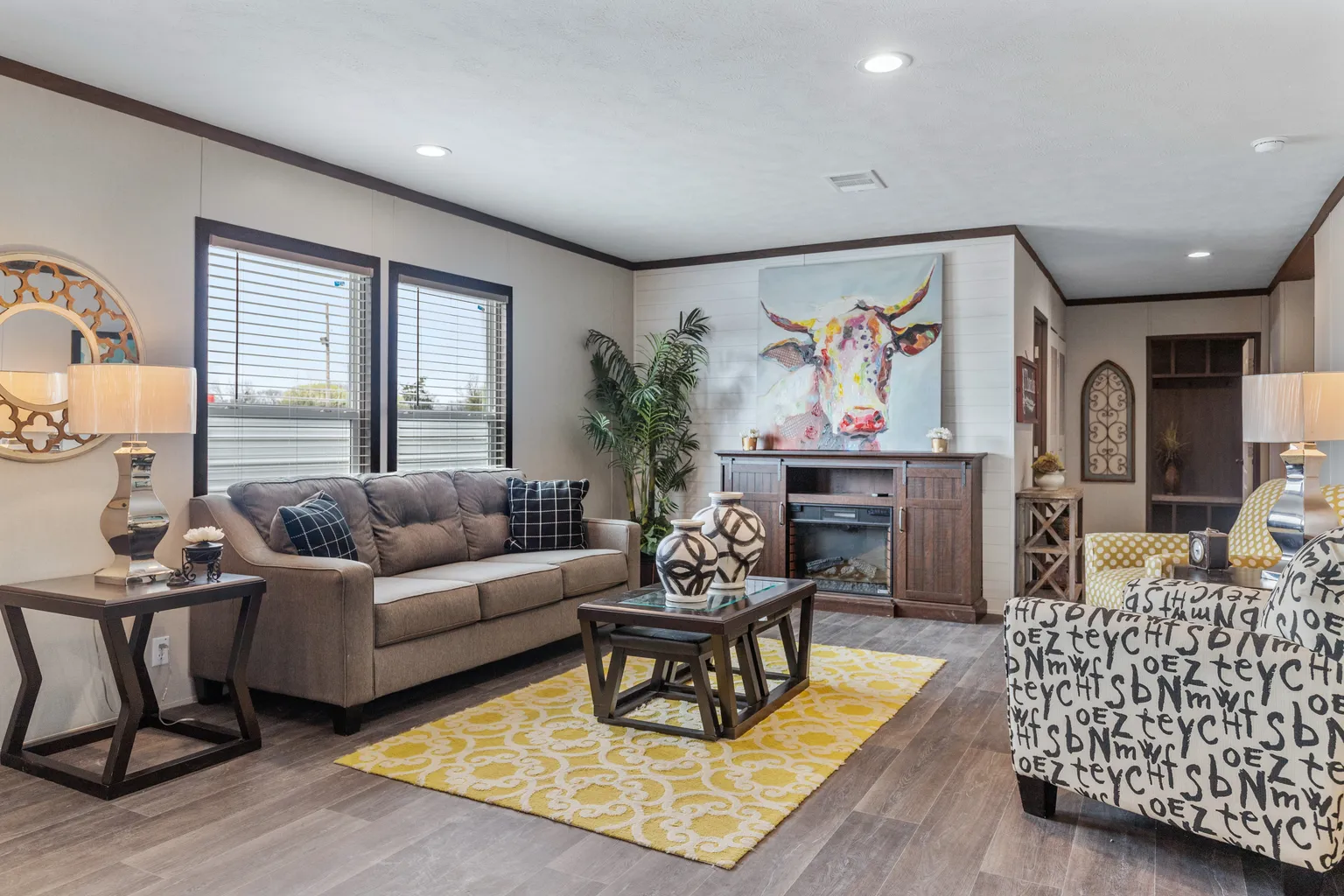 Living room with a brown couch, coffee table and a yellow rug. There’s also other sitting space a painted cow portrait on the entertainment area.