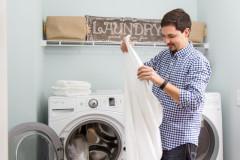 A young man folding a white towel in front of a washer and dryer in a laundry room.
