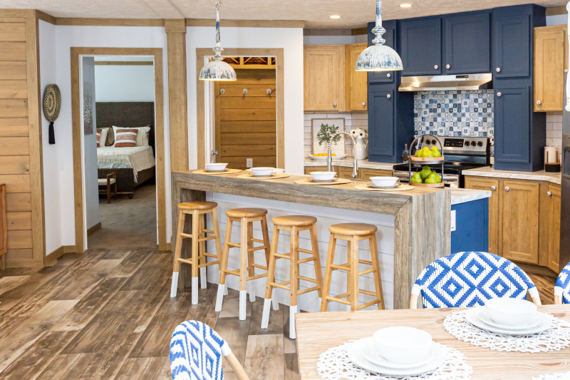 A large kitchen in a manufactured home with blue cabinets, blue ceramic tile backsplash, and a kitchen island with a sit-down bar top with stools.