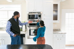 A couple tours a manufactured home kitchen, opening the stainless steel microwave built into white cabinets.