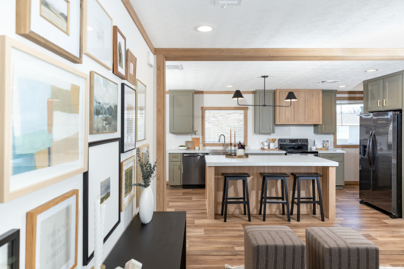 Interior of a manufactured home with a gallery wall to the left and a view of the light wood kitchen island, black appliances and sage green cabinets in the background.