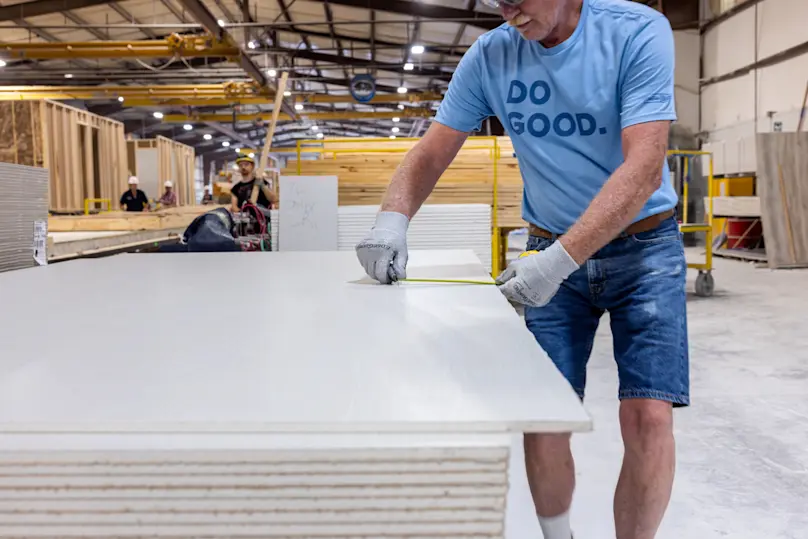 Man in a T-shirt and safety gear measures a sheet of drywall inside a building facility.