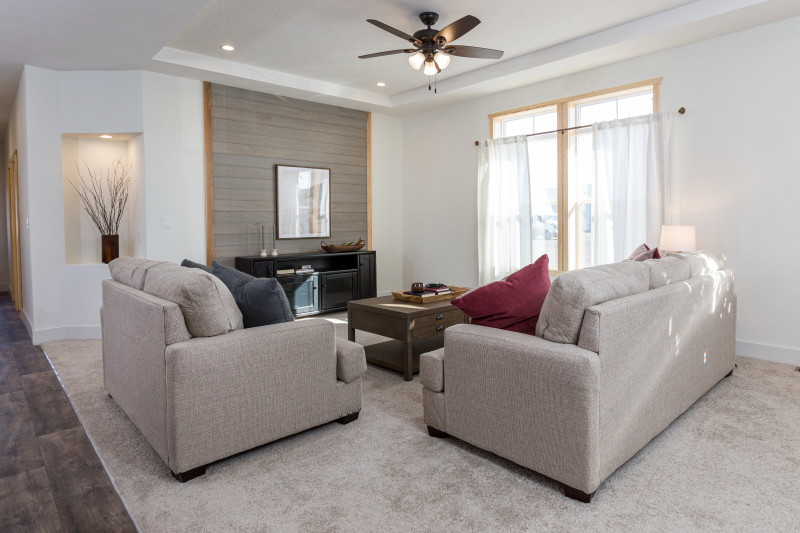There’s a living room with gray and tan accents. There’s a pair of gray couches with a brown coffee table. It also has a gray accent wall, large windows with wood framing, and a small lit cutout for a décor focus.