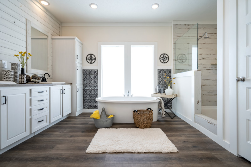 A white rustic bathroom with a soaker tub in the middle of the room in front of two large windows, a walk-in shower on the right and a dual vanity with a shiplap wall on the left.