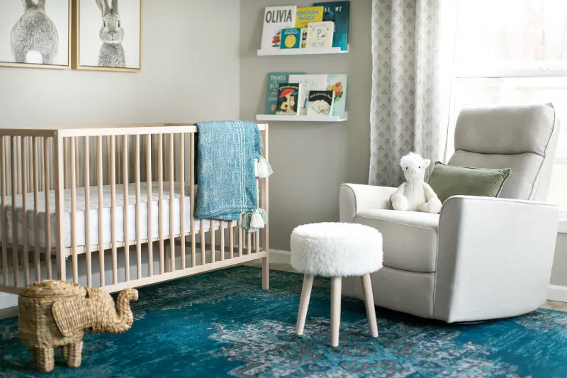 A nursery is decorated with a light wood crib, a blue rug, a white glider chair and shelves with colorful baby books.