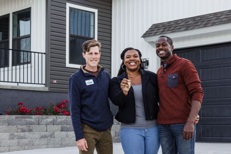 Couple and home center consultant stand and smile in front of a manufactured home, with the woman holding up the key.