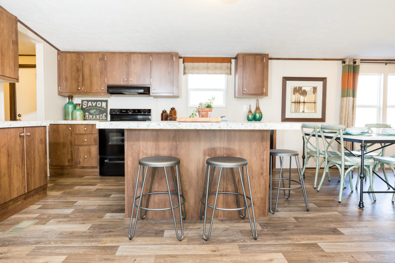 View is of a manufactured home kitchen with a partial section of the dining area. The room has brown cabinets, a kitchen island in the middle, and some silver bar stools around the island. The walls are crème colored and décor is traditional.