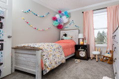 A child’s room is decorated with a flowery duvet, hanging pom poms, pink curtains and a rocking chair with a plush toy sitting in it.