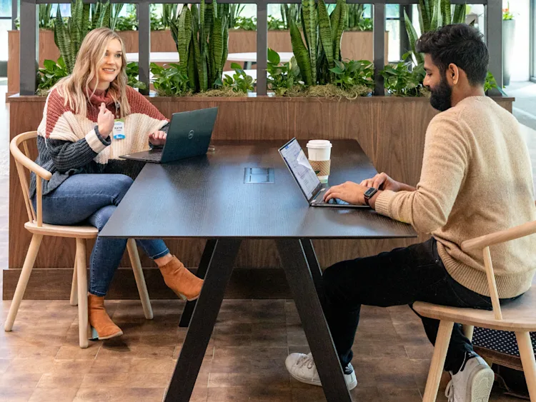 Two team members sit on opposite sides of the table with their laptops open.