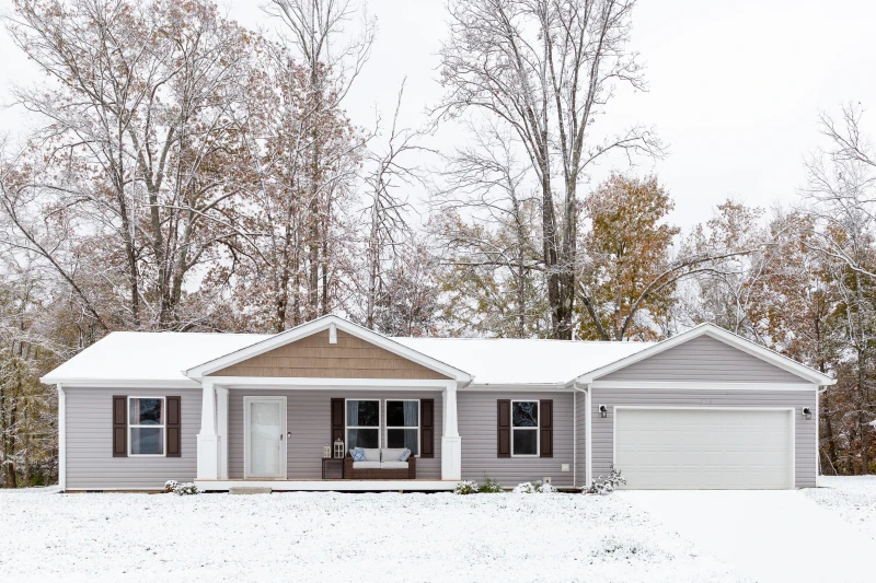 Manufactured home with gray siding, dark brown shutters, white trim and garage with snow covered lawn and roof.