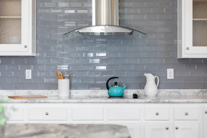 Gray glass backsplash glints in the sunlight in a white cabinet kitchen with a silver hood over it. 