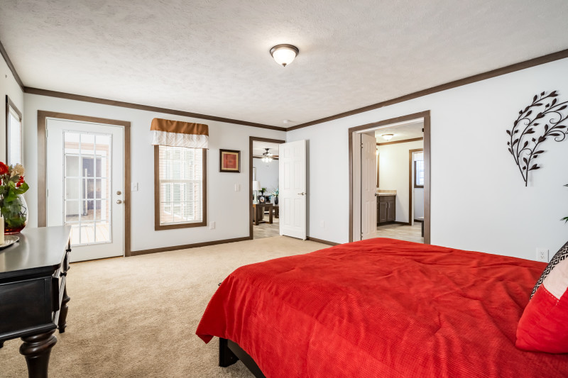 The primary bedroom has wooden molding with white walls. The room has a red bedspread and table. There’s a door out to the deck on one side and a doorway to the primary bath on the other.