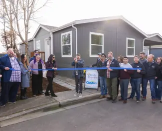 A group of people get ready to cut a ribbon in front of a gray Clayton manufactured home.