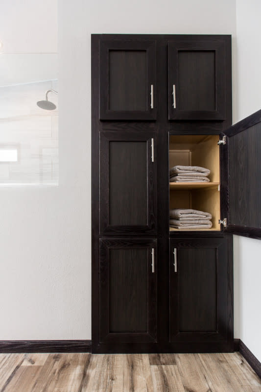 Dark wood linen cupboard with 6 cabinets, one of which is open to show shelf with folded up light brown towels on it.