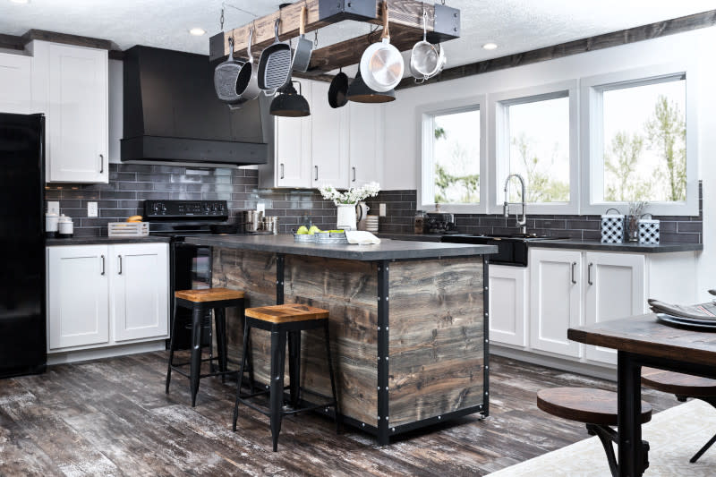 Manufactured home kitchen with kitchen island with a hanging pot rack and breakfast bar, gray title backsplash, white cabinets, black appliances and range hood and 3 picture windows.