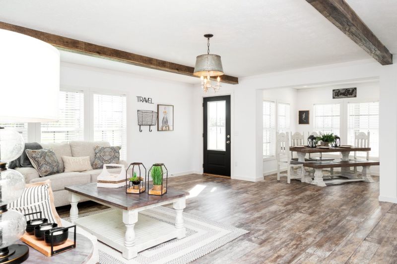 Farmhouse style living room with white walls, weather flooring and ceiling beams, and a door and dining room in the background.