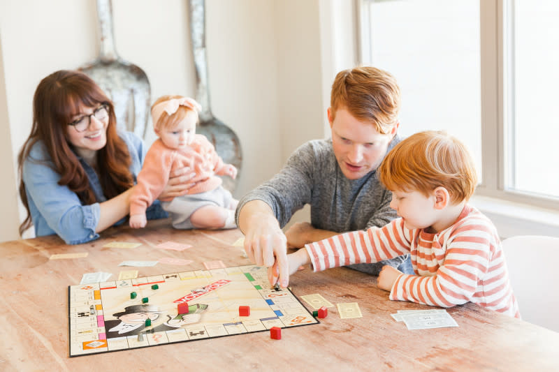 A young family sitting around a Monopoly game while the dad teaches his young son to play.