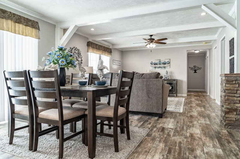 There’s a dining area and living room in view. The area is bright with sunlight and the ceiling has white beams throughout to add height to the rooms. The flooring is a marble looking two tone hardwood. 