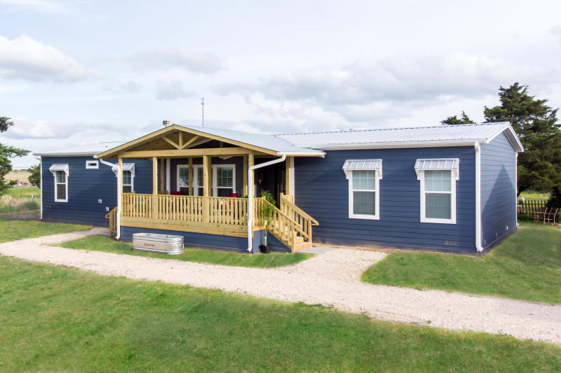 Navy blue ranch style mobile home with covered from porch, plants and metal trough in front yard