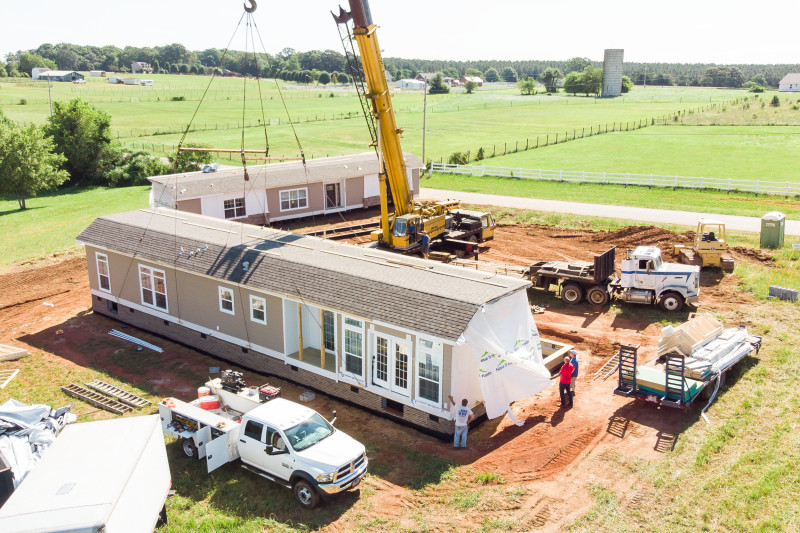 A manufactured home site with 2 home sections with gray-brown siding and white trim, crane, trucks and workers.