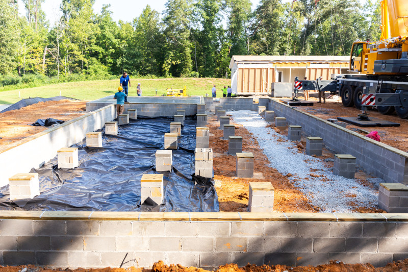 The foundation for a manufactured home is being prepared by workers, with a crane and part of the home in the background to the right.
