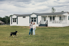 Koven and Josh Bruce standing outside their manufactured home while their dog Lina brings them a stick
