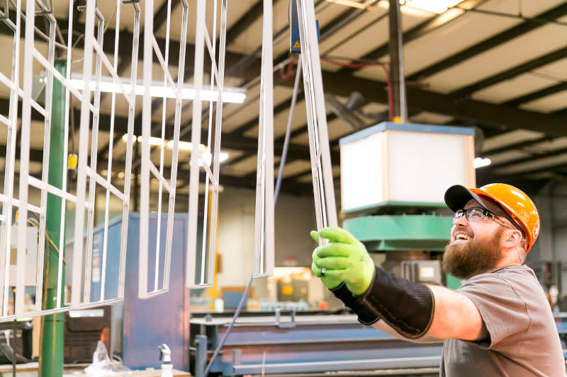 Man wearing hard hat, safety glasses and gloves smiles as he hangs a window frame up on a rack inside of a building facility.